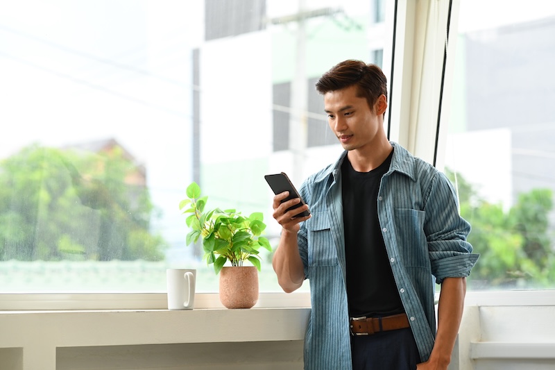Portrait of in asian man in smart casual wear using mobile phone while standing near large window in the creative office.